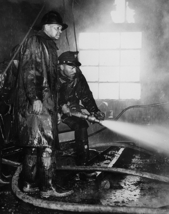 Two black firefighters operate a fire hose in a building